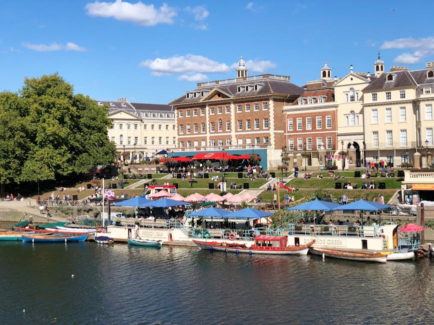 A riverside scene showing a row of boats docked along the water's edge, with a large, mature green tree on the left side. Behind the boats, there are several outdoor seating areas with pink and blue u