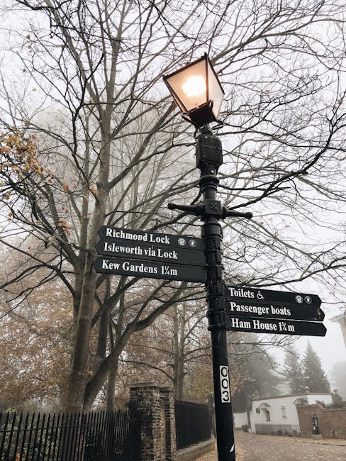 A tall black streetlamp with a lit lantern on top, positioned on a foggy street, surrounded by bare tree branches. Attached to the lamppost are multiple black directional signs with white text indicating various local routes: 'Richmond Lock,' 'Isleworth via Lock,' and 'Kew Gardens 1¼m,' as well as directions for nearby facilities including 'Toilets & Passenger boats,' and 'Ham House 1¼m.' The signs are mounted below the streetlight, visible in the foreground, while a brick fence and a few low buildings are partially visible through the fog in the background. The scene captures a quiet, early-morning atmosphere typical of a house removal area in Richmond upon Thames, with the signs and lamppost supporting navigation for residents and visitors alike, suitable for planning or executing a home relocation or furniture transport in the TW9 postcode area.
