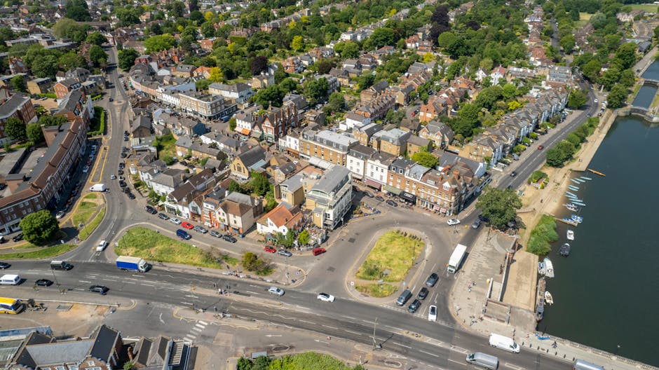 An aerial view of central Richmond upon Thames showing a mix of terraced houses, small commercial buildings, and narrow streets surrounding the river Thames, with boats moored along the waterfront. The image captures a busy intersection near the town centre, where cars and delivery trucks are seen moving along the road, while pedestrians walk on sidewalks. A small parking area with several vehicles is adjacent to the water, and construction or moving activities are not visible in this scene. The dense greenery of trees and residential gardens is evident throughout the neighborhood, with the river on the right side of the image. This setting represents a typical residential area suitable for home relocation by a professional removals service such as [COMPANY_NAME], ensuring efficient furniture transport and packing during house removals in TW9.